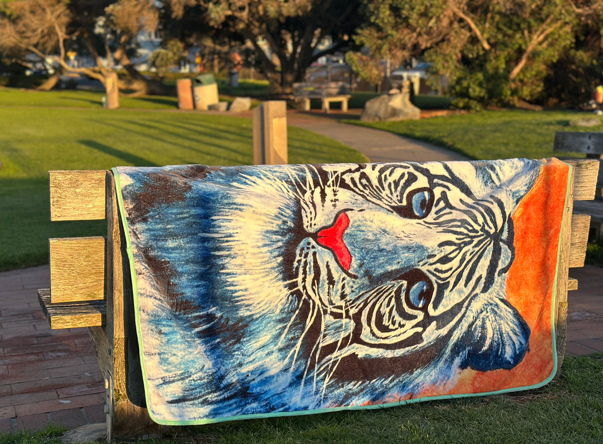 RTEM white tiger flannel throw blanket draped over a wooden park bench in warm afternoon sunlight, displaying the original blue-and-white tiger illustration against an orange background.