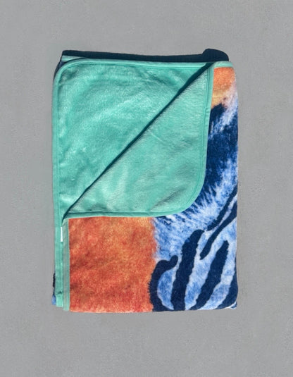 Folded RTEM white tiger throw blanket with one corner turned back to reveal its soft mint-green flannel underside, contrasting with the blue tiger artwork and orange background on the printed front.