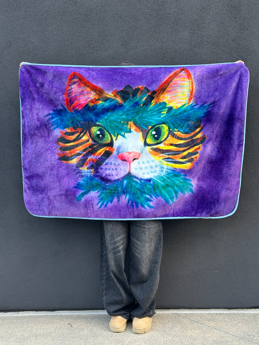 A person holds up RTEM's Healer Cat flannel throw blanket against a dark gray wall, showing the full purple front design with a colorful cat, bright green eyes, orange-striped cheeks, a pink nose, and turquoise-green magical herbs gathered around its head and mouth.
