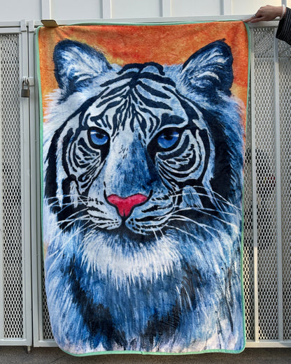 Full front view of RTEM's white tiger flannel throw blanket hanging against a white metal fence, displaying a blue-eyed white tiger with black stripes, a pink nose, an orange background, and mint-green trim.