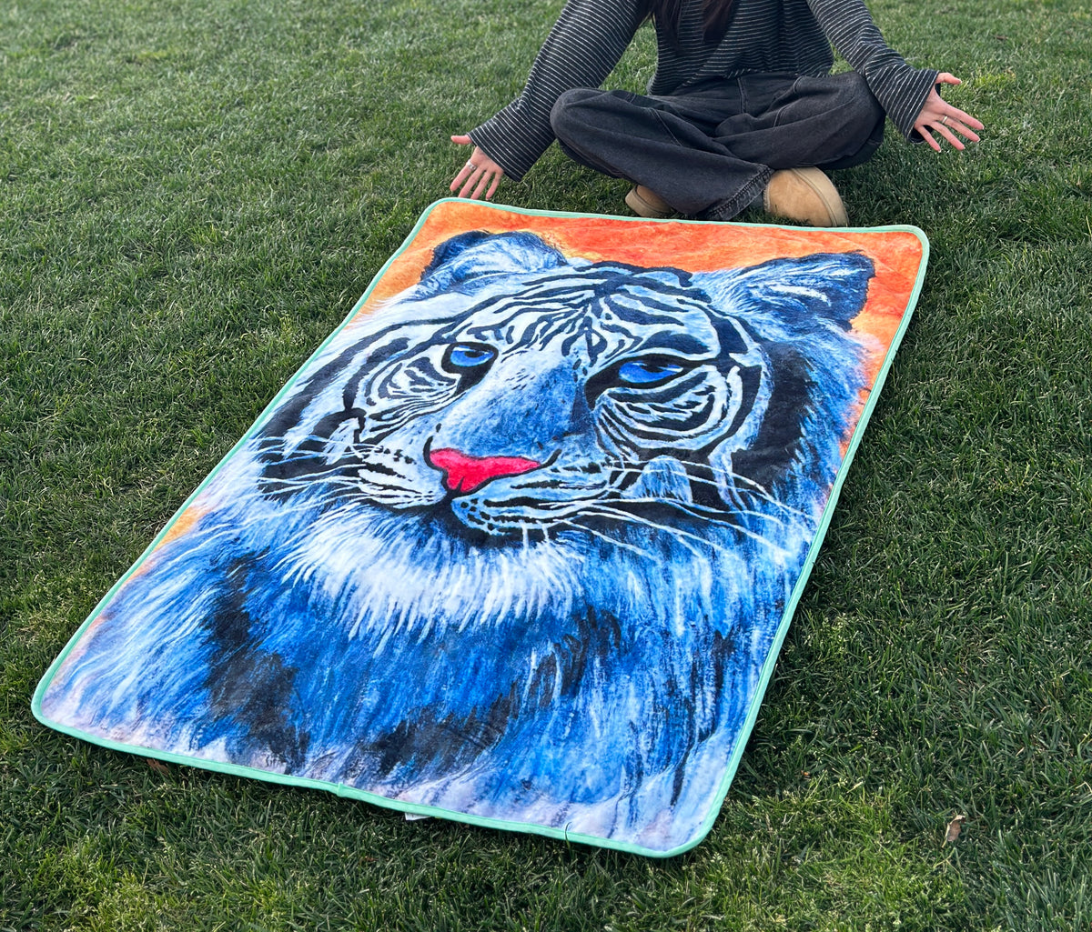 RTEM's white tiger flannel throw blanket laid flat on green grass, with a person sitting cross-legged behind it. The blanket features an original blue-eyed white tiger illustration with bold black stripes, a pink nose, an orange background, and mint-green edging.
