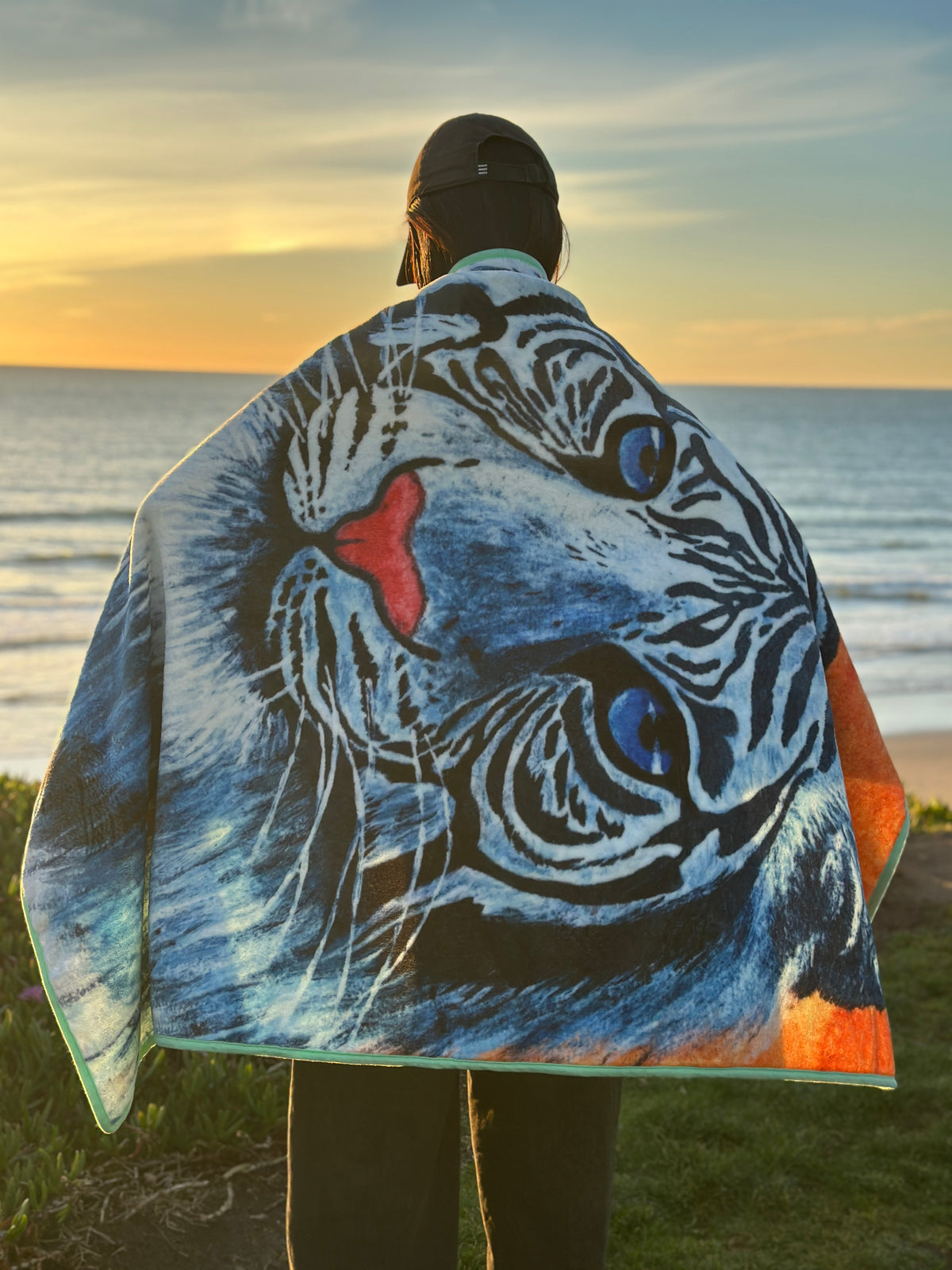 A person wrapped in RTEM's original white tiger flannel throw blanket, featuring a blue-eyed white tiger with black stripes and a pink nose, standing by the ocean at sunset.