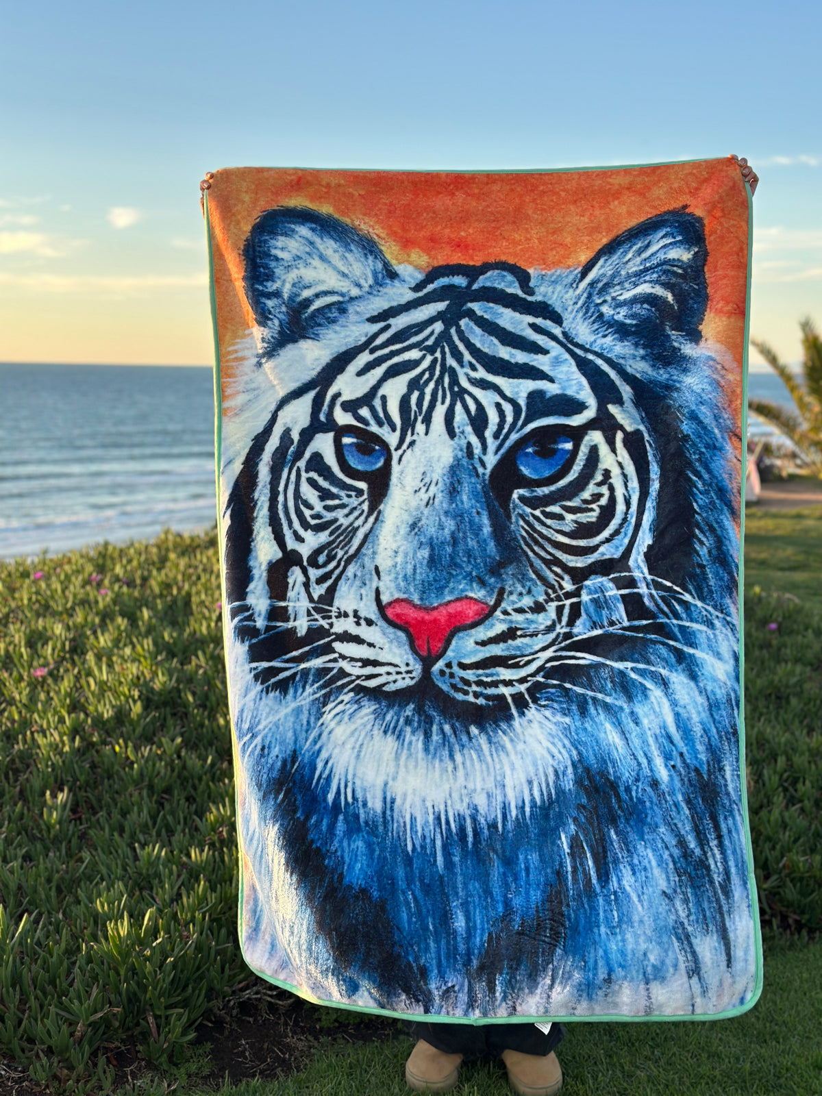 RTEM white tiger throw blanket held vertically beside the ocean, featuring an original illustration of a blue-eyed white tiger with bold black stripes and a pink nose on an orange background.