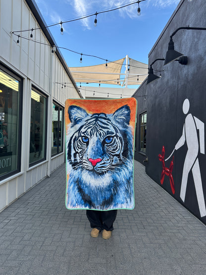 A person holding RTEM's white tiger throw blanket upright in an outdoor walkway beneath string lights, showing the full blue-eyed white tiger with pink nose on an orange background.